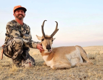 A hunter kneeling next to a pronghorn antelope on open Wyoming plains, wearing a camouflage jacket and orange hat, showcasing their successful hunt with SNS Outfitter & Guides.